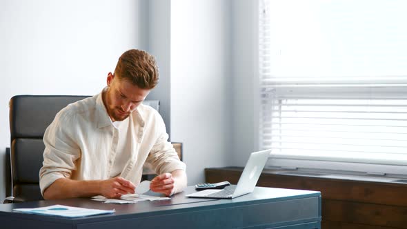 Bearded guy manager in yellow shirt sorts different paper receipts on grey table alt