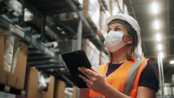 Warehouse Worker Wearing Medical Mask Safety Vest and Hardhat Standing Near Shelves with Goods Using alt