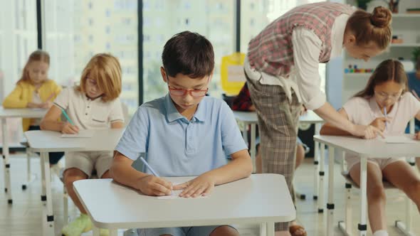 Asian Boy Pupil Wearing Glasses Sits at a First Desk in the Classroom and Fills Out Tests and the alt