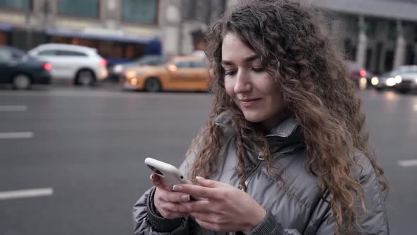 Young Female City Dweller Is Using Smartphone Standing Near Road in Town Viewing Social Nets and alt