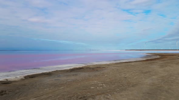 Drone Flight Over Smooth Surface of Colorful Pink Lake at Cloudy Sunrise with Wind Farm on alt