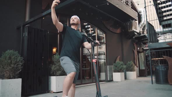 Young Man Standing with Escooter in City Downtown and Taking Selfie with Smartphone alt