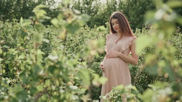 Young pregnant woman with flowing hair in a pink summer dress stands among the green plants.
