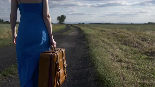 Girl in blue dress with suitcase on country road in summer.