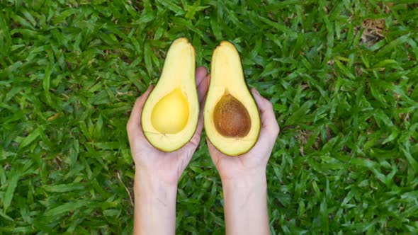 Woman Hands Hold and Open Two Cut Halves of Fresh Green Avocado on Green Grass alt