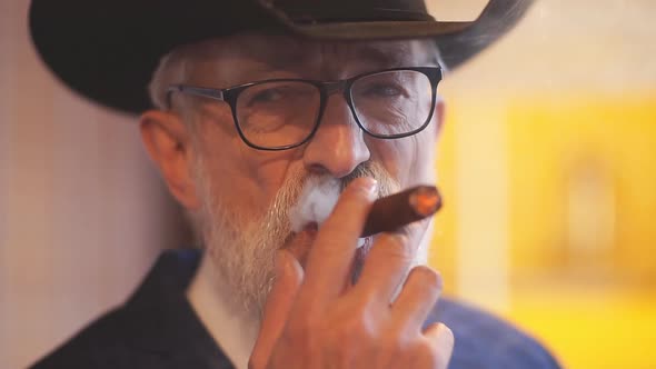 Stylish Old Man in Wide Brimmed Hat and Rich Blue Mens Suit Smoking Cigar Indoor alt
