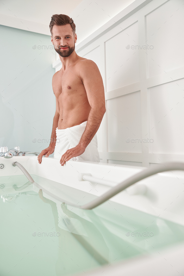 Attractive man with bare chest stands near hydro massage tub in spa salon