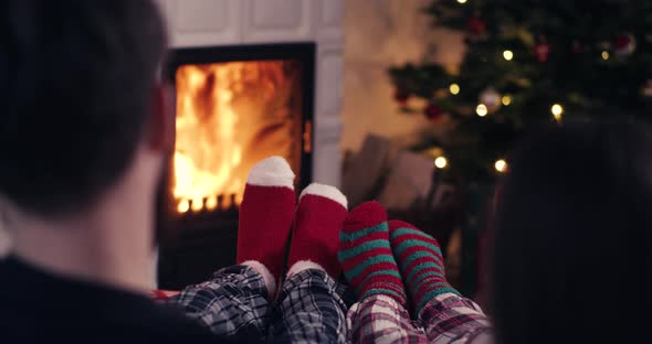 Couple feet in christmas woolen socks near fireplace with decorated tree in backg alt