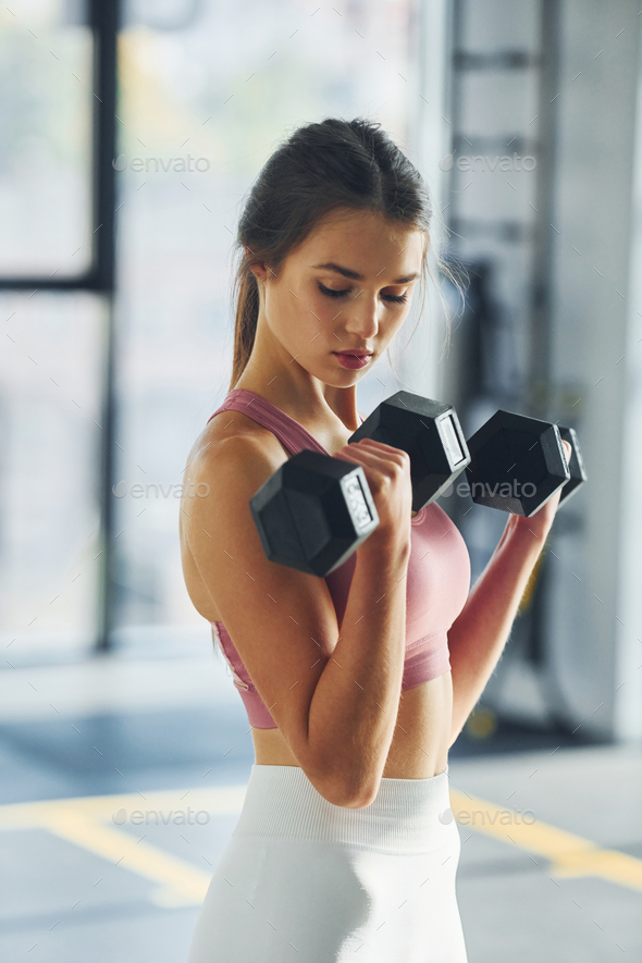 Holding dumbbells. Beautiful young woman with slim body type is in