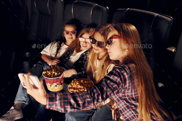 Holding phone in hand. Group of kids sitting in cinema and watching movie together