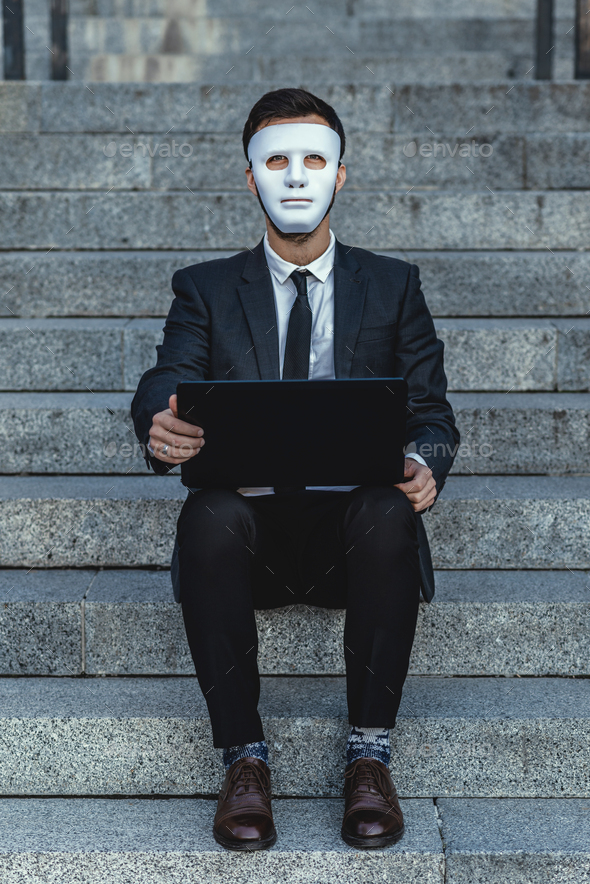 Businessman with a face mask and a laptop looks straight. Sits on the steps of a business building