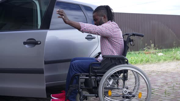 Disabled African American Man Riding Wheelchair to Car Making Effort Sitting on Driver's Seat alt