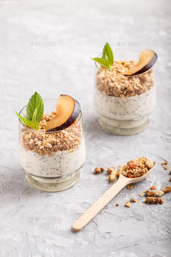 Yoghurt with plum, chia seeds and granola in a glass and wooden spoon on gray concrete