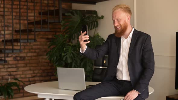 Cheerful Redhead Bearded Businessman Making Video Call on the Smartphone Waving