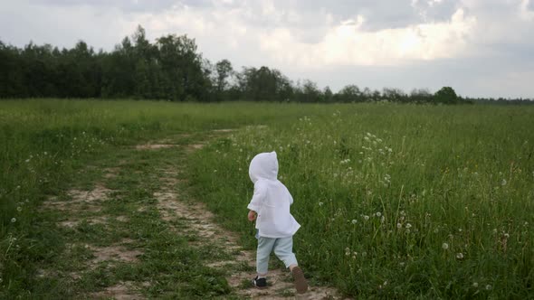 Boy in Blue Pants and a White Jacket in a Hood Runs Along the Road alt