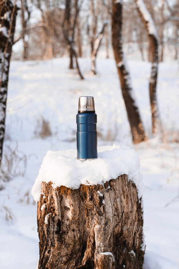 Blue thermos standing on a snow-covered stump. Camping vacuum flask in the winter forest during the