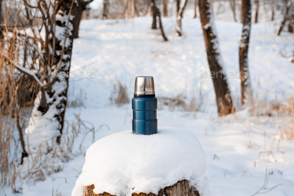 Blue steel thermos standing on a snow-covered stump. Camping vacuum flask in the winter forest