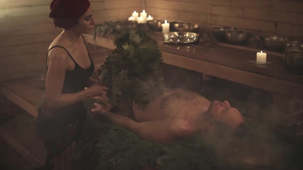 A woman conducts a bathing ritual for a man with the help of brooms. A man lies on spruce branches.