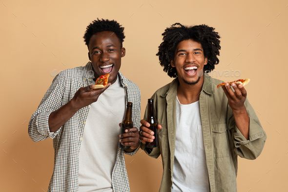 Men\'s Leisure Concept. Two Black Guys Holding Beer Bottles And Eating Pizza