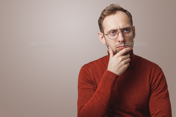 Young thougthful man holds chin and looks pensively away wears spectacles and red turtleneck