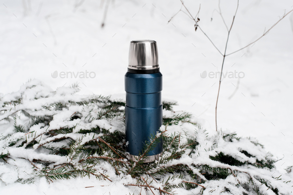 Thermos standing on pine branch in the snow. Vacuum flask outdoors. Winter camping in the forest