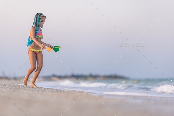 A girl with african braids in a summer costume plays on the beach with  shells near the sea with Stock Photo by YouraPechkin