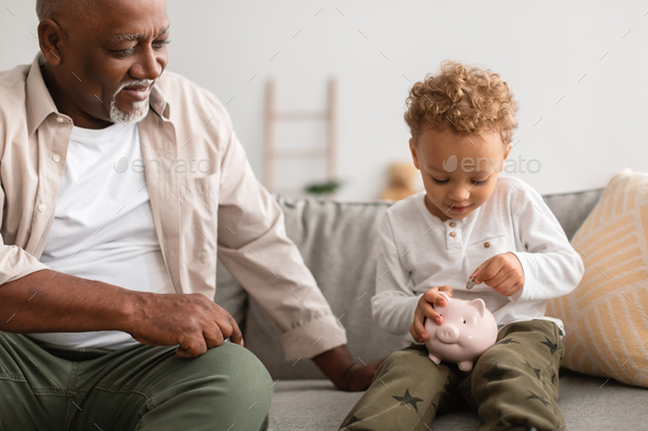 African American Grandfather And Grandson Putting Money In Piggybank Indoor