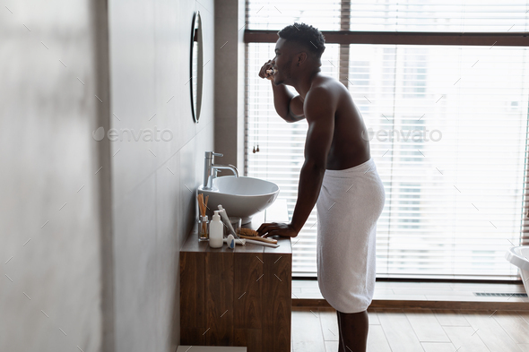 African american guy brushing teeth standing near basin in bathroom