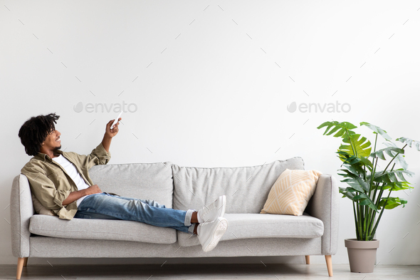 Home Air Conditioning. Black Guy Using Remote Controller While Relaxing On Couch