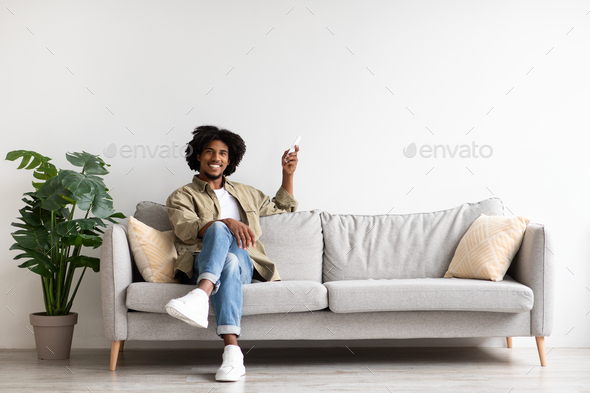 Home Comfort. Young Black Man Sitting On Couch And Holding Remote Controller