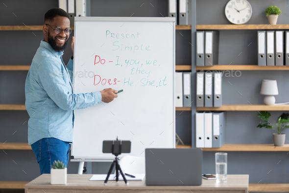 Happy millennial attractive black man tutor points to English rules on blackboard, looks at phone