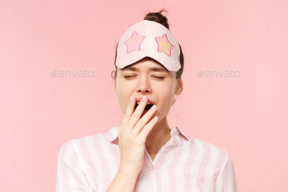 Woman in pyjama shirt and sleeping mask yawning and covering mouth, wanting to sleep