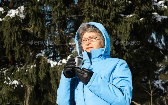 Elderly woman with glasses holding mug of hot drink outside. Female pensioner in blue ski jacket