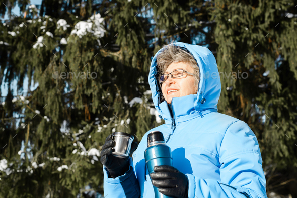 Relaxing elderly woman with glasses holding thermos and mug with hot drinks outdoors. Pensioner in