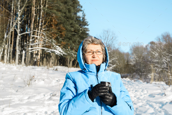 Smiling elderly woman with glasses holding mug with hot drink outside. Female pensioner in blue ski
