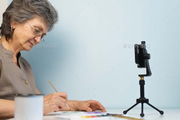 Online drawing workshop. Senior woman in glasses painting with watercolors indoors. Retired hobby
