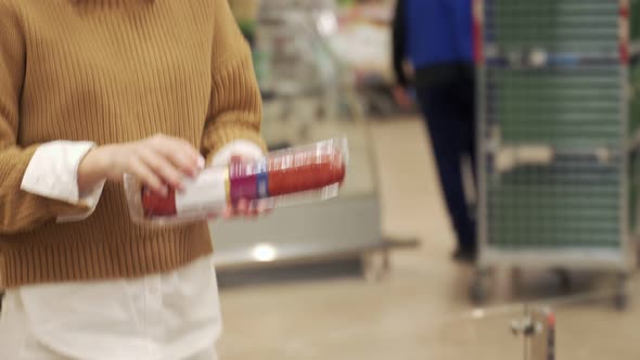 Young Woman Takes a Sausage From a Supermarket Shelf Holds a Smartphone Scans the Barcode on the alt