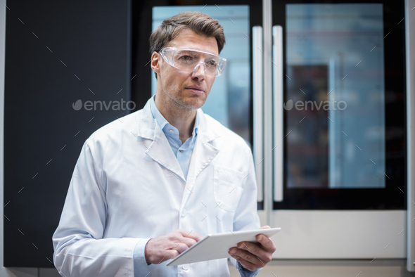 Portrait of man wearing lab coat and safety goggles holding tablet at machine