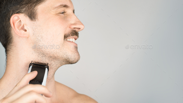 Young smiling man cutting stubble. Mustachioed man with modern electric beard trimmer. Close-up