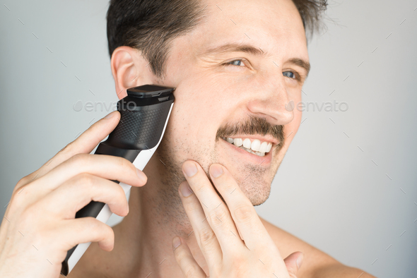 Young smiling man cutting stubble, close-up. Man with mustache and modern electric beard trimmer