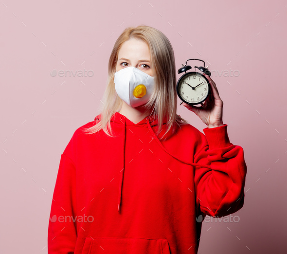 Woman in FFP2 anti-dust standart face mask hold alarm clock on pink background