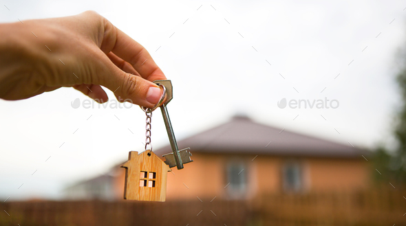 Hand with a key and a wooden key ring-house. Background of fence and cottage