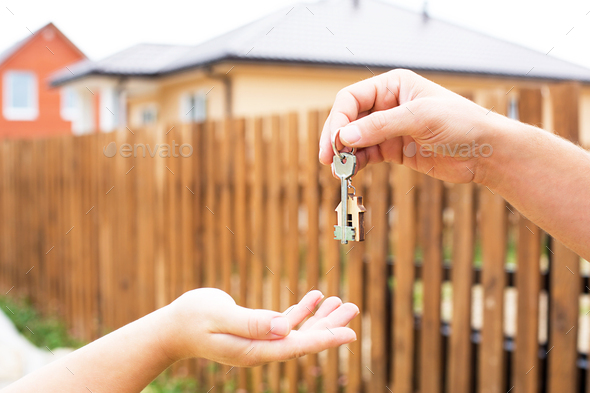 Hand with a key and a wooden key ring-house. Background of fence and cottage.
