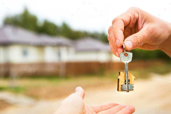 Hand with a key and a wooden key ring-house. Background of fence and cottage