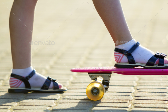 Child slim legs in white socks and black sandals on plastic pink skateboard on bright sunny summer