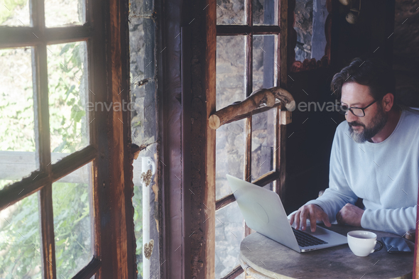 Mature man at work on laptop computer sitting at the table inside a home cozy cabin