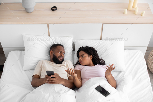 Angry sad millennial black woman gesturing and look at husband for addiction to phone, online game