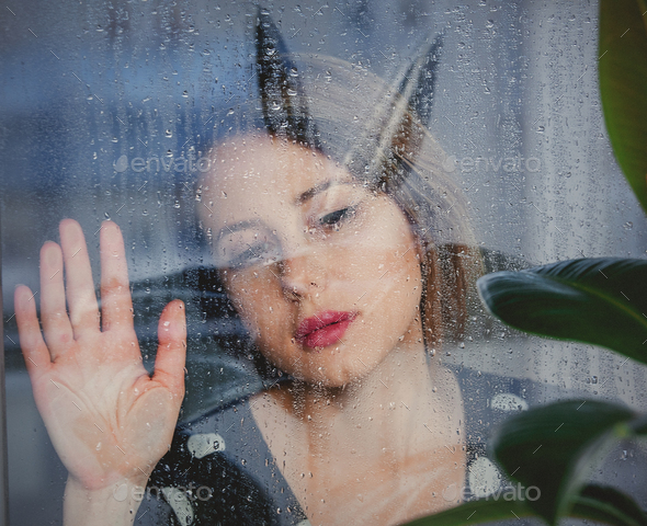 young sad woman near wet window after the rain misses the ficus plant