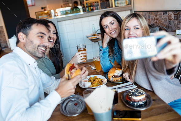 Happy group of friends taking selfie with smart phone while eating burgers in a restaurant.
