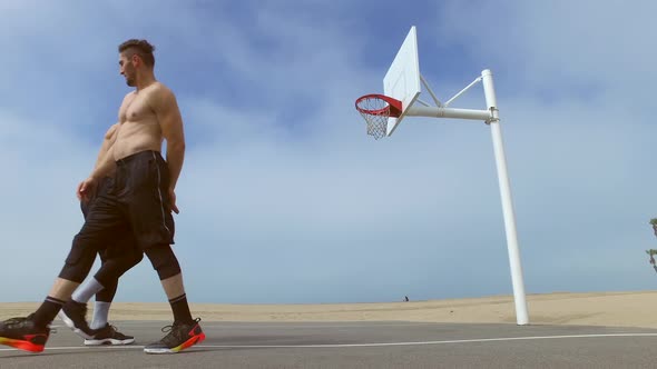 Two men play one-on-one basketball hoops on a beach court. alt